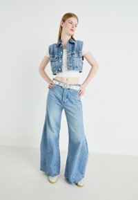 Farmerdzseki, gombos záródással, rövidített fazonban, széles lábú világoskék farmernadrággal kombinálva. Fehér öv fémes részletekkel. A modell egy sima háttér előtt áll.