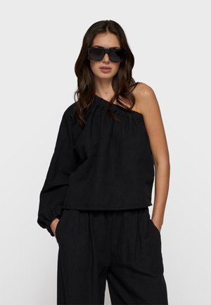 Frau mit übergroßen schwarzen Sonnenbrillen, schwarzem, einseitigem langärmligem Oberteil und weitem schwarzen Hosen, die mit den Händen in den Taschen vor weißem Hintergrund steht.