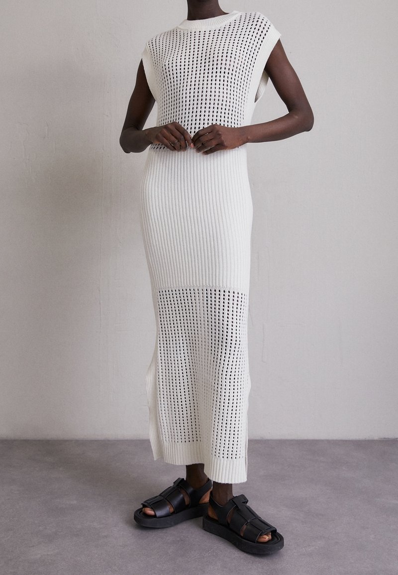 Weißes gestricktes Kleid mit geripptem Muster und offenem Design. Mit ärmellosem Schnitt und seitlichen Schlitzen. Kombiniert mit schwarzen Sandalen.