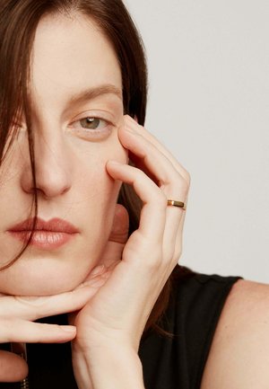 Anillo de oro en un dedo apoyado en una barbilla, con una vista en primer plano de un rostro de piel clara, maquillaje sutil y cabello castaño parcialmente visible.