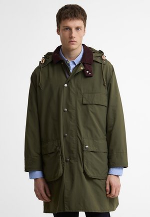 Jeune homme portant un parka à capuche vert olive avec des boutons-pression et de grandes poches avant, par-dessus une chemise bleu clair.