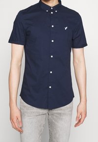 Uomo che indossa una camicia a maniche corte blu navy adatta, con bottoni bianchi e un piccolo logo di un uccello ricamato sul petto.