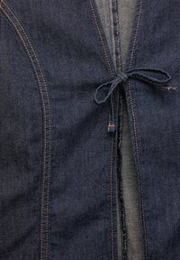 Mörkblå denimplagg med mittknutstängning. Har synlig sömnad, en strukturerad yta och en öppen halsringning.
