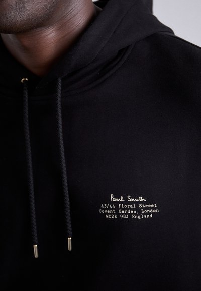 Sudadera negra con capucha que presenta un cordón con extremos dorados. Logo bordado y dirección en blanco en la parte delantera. Textura de tela suave.