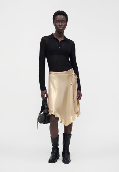 Donna che indossa una maglia nera a maniche lunghe con collo, gonna portafoglio in raso dorato con volant, stivali neri, tiene una borsa nera, in piedi su sfondo bianco.