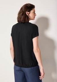 Blouse noire à manches courtes avec une coupe décontractée, ourlet élastique et un pli central au dos, assortie à un pantalon bleu. Tissu lisse et léger.