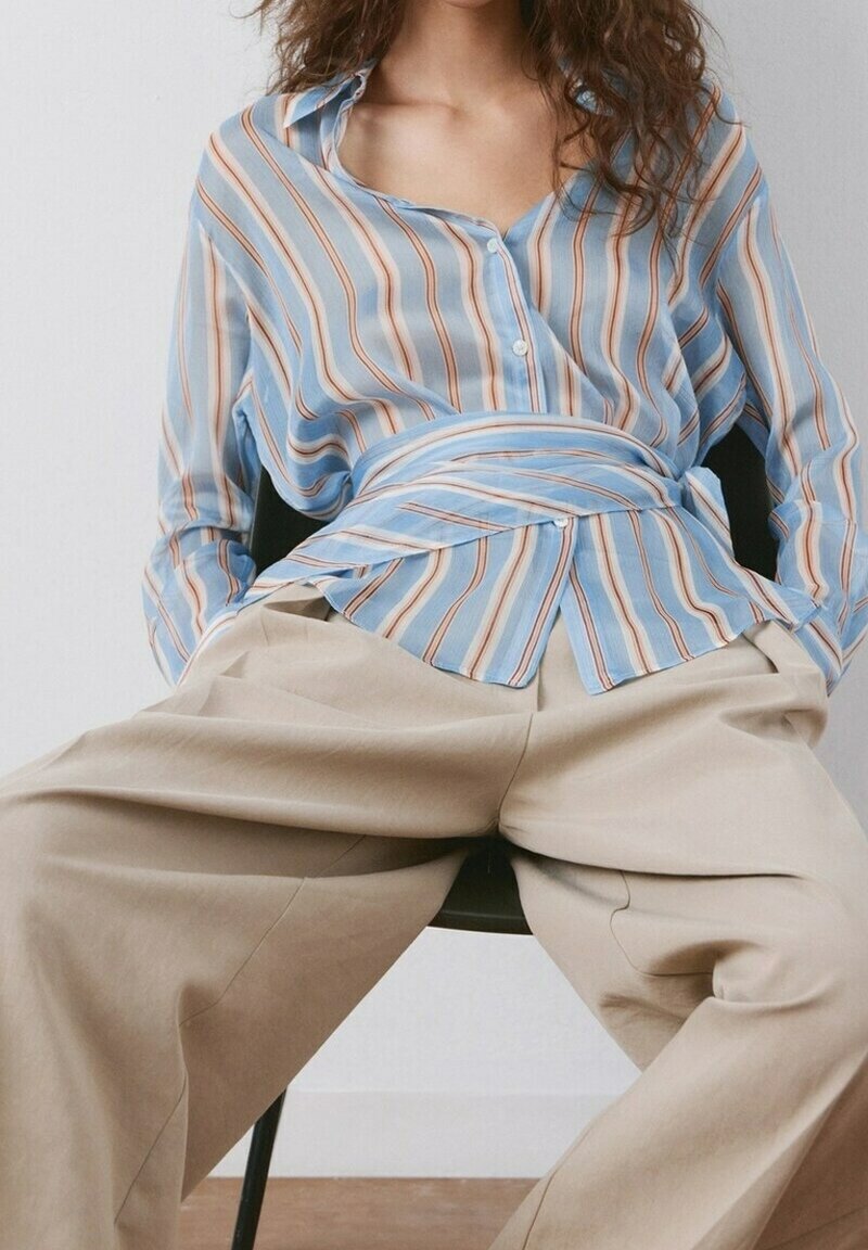 Personne assise portant une chemise à rayures bleues boutonnée avec un détail enveloppant et un pantalon ample beige à jambes larges sur un fond uni.