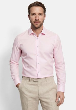 Homme portant une chemise rose clair à boutons et un pantalon beige, debout avec une main dans la poche devant un fond uni.