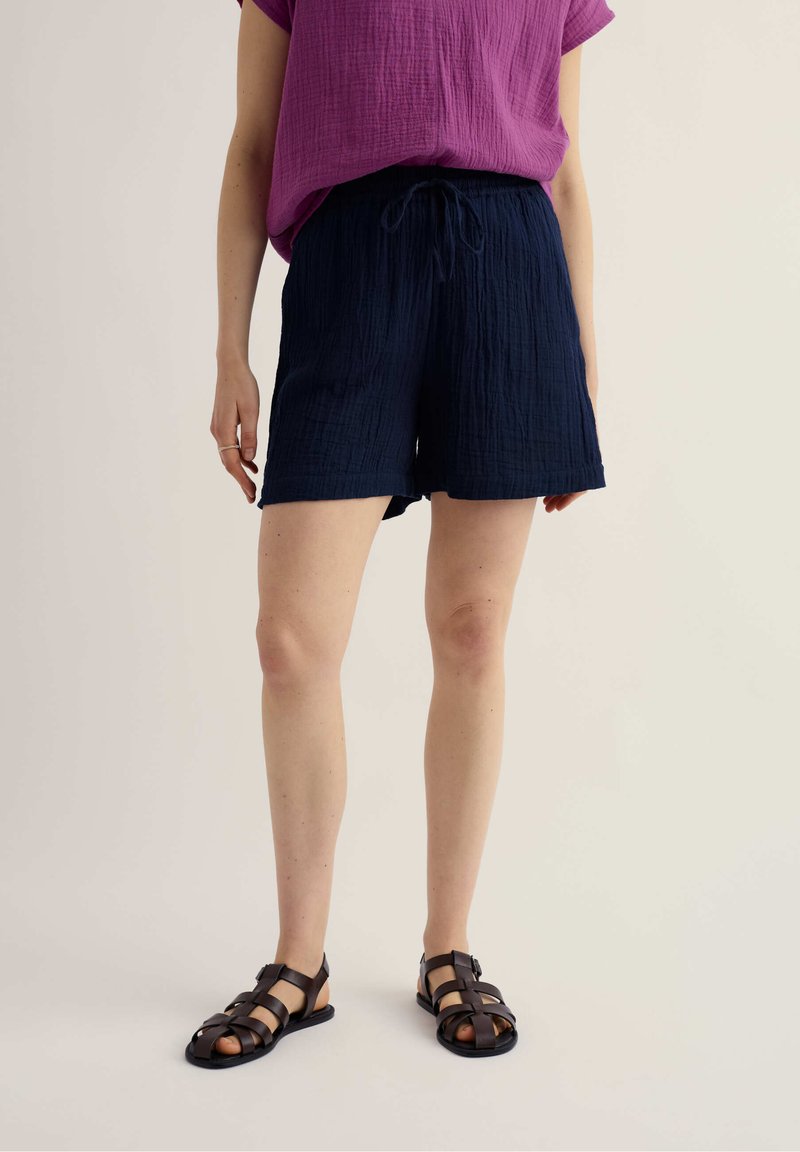 Person trägt ein lila kurzärmeliges Oberteil, marineblaue Shorts mit Kordelzug und dunkelbraune Sandalen mit Riemchen, steht vor einem hellen Hintergrund.
