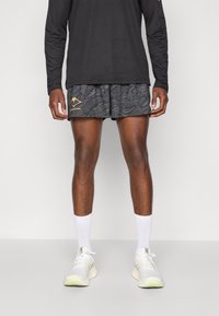 Black long-sleeve shirt, patterned gray shorts with a gold mountain logo, white ankle socks, and white sneakers with green accents.