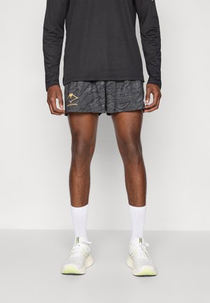 Schwarzes Langarmshirt, gemusterte graue Shorts mit einem goldenen Berglogo, weiße Knöchelsocken und weiße Sneakers mit grünen Akzenten.