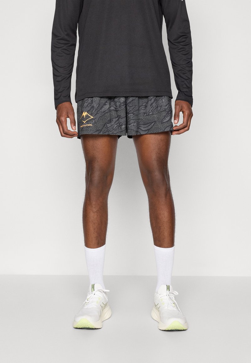 Black long-sleeve shirt, patterned gray shorts with a gold mountain logo, white ankle socks, and white sneakers with green accents.