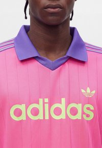 Άτομο που φοράει ροζ μπλούζα Adidas με μωβ γιακά και κίτρινο λογότυπο και γράμματα Adidas στο στήθος, κοντινό πλάνο από τον λαιμό μέχρι τον άνω κορμό.