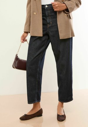 Femeie purtând blugi cu talie înaltă, de culoare albastru închis, sacou maro cu carouri, pantofi maro și ținând o geantă mică maro cu o curea aurie.