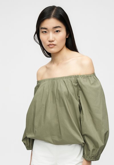 Νεαρή γυναίκα με μακριά σκουρόχρωμα μαλλιά, φορώντας μια χαλαρή ελιάς πράσινη μπλούζα με το ένα ώμο έξω και λευκό κάτω μέρος, μπροστά από ένα απλό φόντο.