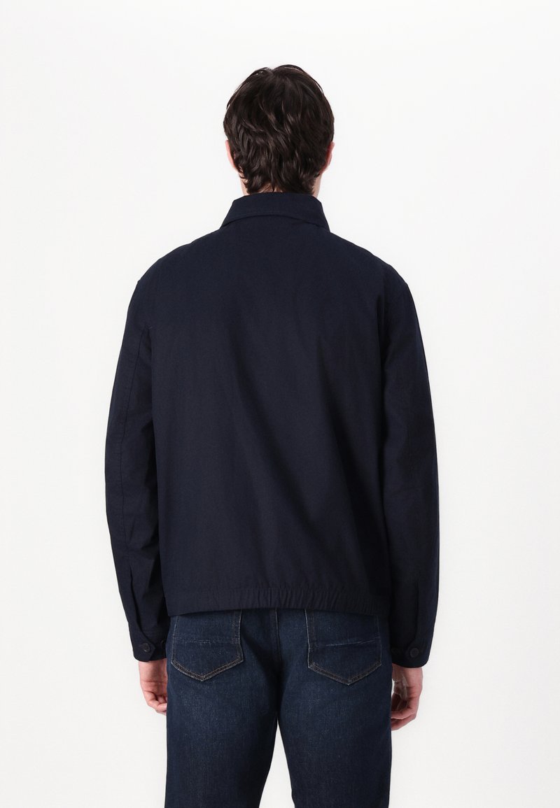 Marineblaue Jacke mit Kragen und elastischem Bund, ausgestattet mit langen Ärmeln und einer glatten Stoffoberfläche. Getragen mit blauen Jeans.