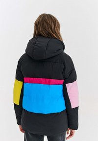 Osoba obrátená chrbtom, oblečená v čiernej pufrovanej bunde s kapucňou, so širokými farebnými blokmi v ružovej, modrej, žltej a čiernej na rukávoch.