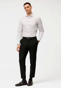 Weißes Hemd mit kleinen schwarzen Mustern, schwarze Hose und dunkle Schuhe. Das Outfit verfügt über einen Button-Down-Kragen und eine schlanke Passform.