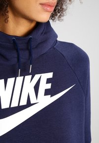 Navy blue hoodie with a high collar, featuring a large white Nike logo and silver hardware for drawstrings. Soft fabric with a relaxed fit.