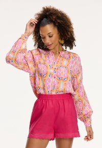 Krāsains blūze ar rakstu rozā, oranžā un zilā krāsā, ar pogām un garām piedurknēm, savienota ar rozā šortiem ar viļņotām malām.