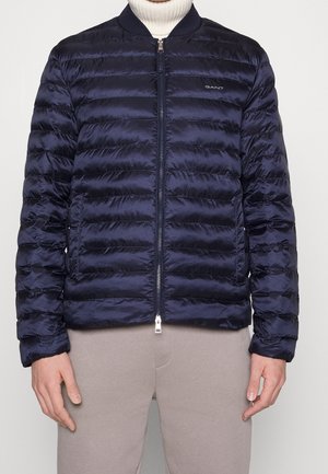 Mørkeblå, quiltet jakke med lynlås og vandret syning, båret over en hvid rullekrave og beige bukser, forfra med hænderne afslappet.