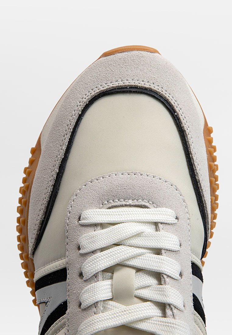 Zapatilla deportiva con parte superior de ante beige y gris, detalles en negro, cordones blancos y suela de goma texturizada color caramelo.