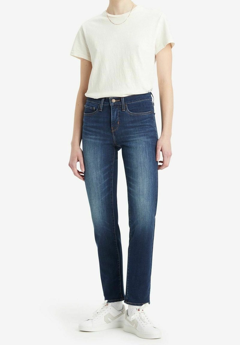 Jeans ajustados azul oscuro de tiro alto, combinados con una camiseta de color claro y zapatillas blancas. Los jeans presentan un diseño clásico de cinco bolsillos.