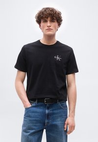 Νεαρός άνδρας με σγουρά μαλλιά, φορώντας μαύρο t-shirt Calvin Klein και μπλε τζιν, στέκεται μπροστά σε απλό ανοιχτό φόντο.