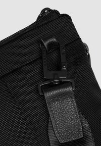 Detail einer schwarzen Nylon-Tasche mit einem strukturierten Tragegurt, Kunststoffbeschlägen und einem Reißverschluss. Der Stoff hat ein geripptes Muster und glatte Lederelemente.