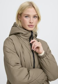 Gepolsterte, khakifarbene Pufferjacke mit hohem Kragen, schwarzem Reißverschluss und elastischen Bündchen. Verfügt über eine glatte Textur und eine taillierte Silhouette.
