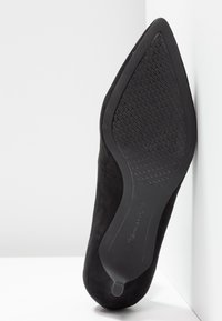 Semelle de chaussure noire à bout pointu avec grip en caoutchouc texturé et marque Tamaris, taille 37, reposant contre un coin blanc.
