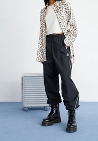 Persona con chaqueta de estampado de leopardo, top corto blanco, pantalones de chándal negros de Adidas con franja verde y botas de combate negras de plataforma cerca de una maleta plateada.