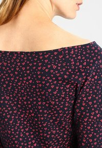 Mujer de piel clara y cabello castaño claro lleva una blusa azul marino sin hombros con un estampado de corazones rojos.