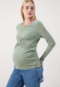 Hosszú ujjú, bordás várandós blúz világoszöld színben, csavart elülső résszel. Lazán illeszkedő kék farmernadrággal párosítva.