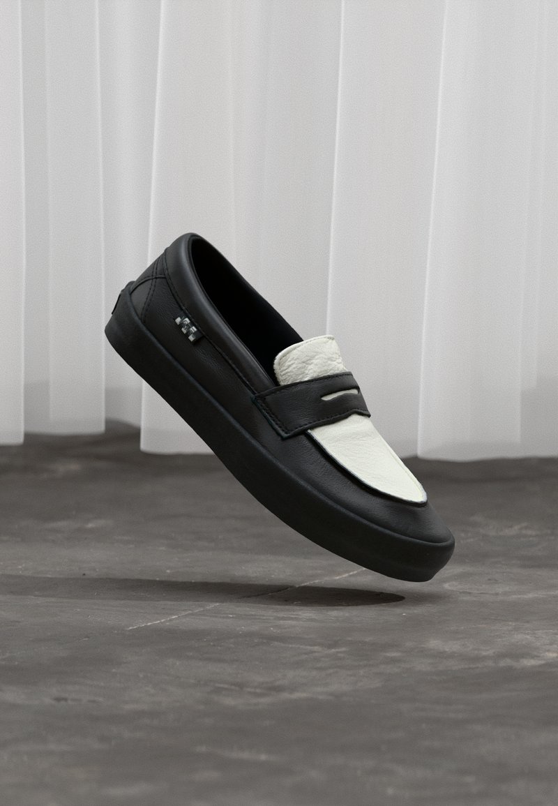 Schwarz-weißer Leder-Slipper mit dicker Sohle, der über einem grauen Betonboden vor durchsichtigen weißen Vorhängen schwebt.