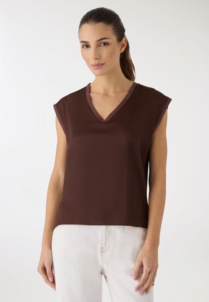 Donna con capelli scuri raccolti in una coda di cavallo, che indossa un top senza maniche color marrone scuro con scollo a V e pantaloni chiari, su uno sfondo semplice.