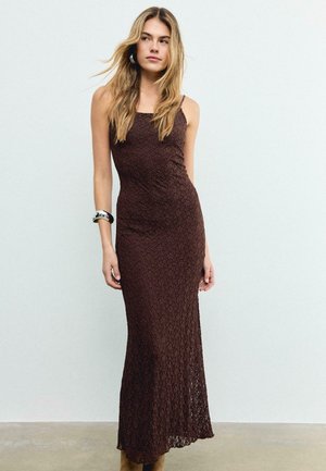 Kvinde med langt blondt hår iført en ærmeløs, mørkebrun maksidragt med tekstur og sølvarmbånd, stående mod en ensfarvet lys væg.