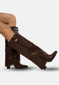 Botas de cuero marrón hasta el muslo con punta afilada, tacón bajo y con una cremallera visible. Presentan un amplio puño y acentos metálicos.