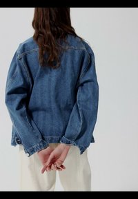 Osoba s dlhými hnedými vlasmi, oblečená v modrej rifľovej bunde a krémových nohaviciach, stojaca so spojenými rukami za chrbtom.