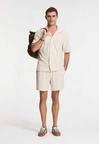 Cămașă crem deschis cu mâneci scurte și pantaloni scurți asortați, confecționate dintr-un material texturat, purtate cu adidași maro. Un rucsac verde este cocoțat pe un umăr.