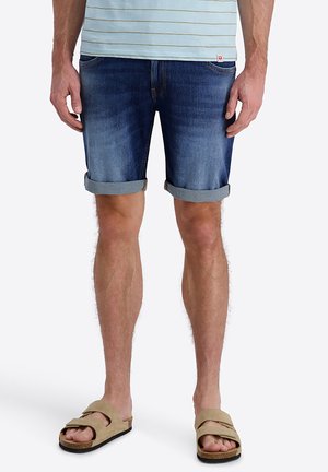 Mann trägt hochgekrempelte blaue Denim-Shorts, gestreiftes Hemd und beige Schlupfsandalen, steht vor einem einfarbigen Hintergrund.