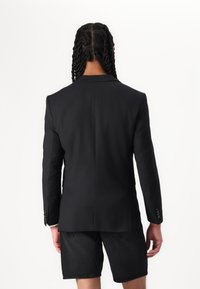 Sort blazer med en slank pasform, klapkraver og knapmanchetter. Har en glat tekstur og rene linjer, båret over mørke shorts.