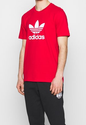 Osoba nosząca czerwoną koszulkę Adidas z białym logo oraz czarne spodnie z mniejszym białym logo Adidas na lewym udzie.