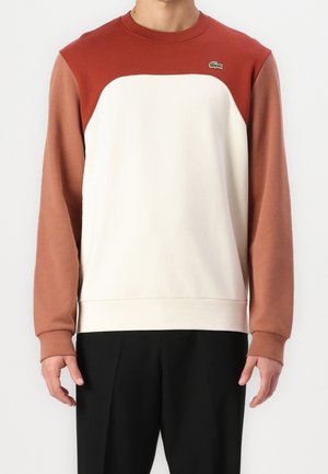 Homme portant un sweat-shirt bicolore avec un devant blanc, des épaules rouge rouille et des manches marron clair, associé à un pantalon noir.
