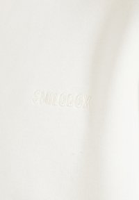 Krämfärgad bomulls-tröja med strukturväv och broderad "SMILODOX"-logotyp i matchande tråd på framsidan.