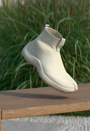 Witte sneaker in sokstijl met gespleten teen en dikke zool zwevend boven een houten bankje, met een wazige groene grasachtergrond.