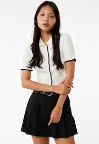 Krótkosleeve'owa, prążkowana, biała bluzka z czarnymi wykończeniami i guzikami, zestawiona z czarną spódnicą typu skater oraz czarnym paskiem ze srebrną klamrą.