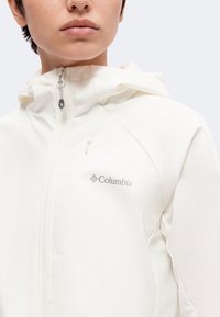 Νεαρό άτομο που φοράει λευκό μπουφάν Columbia με κουκούλα και μπροστινό φερμουάρ, που δείχνει το λογότυπο της μάρκας στο στήθος.