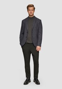 Marineblauer strukturierter Blazer mit zwei Knöpfen, grauem Rollkragenpullover, dunkler Hose und schwarzen Schnürstiefeln. Das Model steht aufrecht vor einem neutralen Hintergrund.