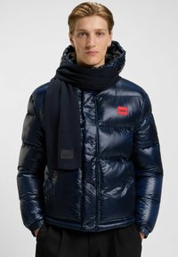 Giacca puffer blu navy con finitura lucida, cappuccio, patch logo rosso e una sciarpa nera abbinata con etichetta di marca. Strutturalmente morbida e calda.
